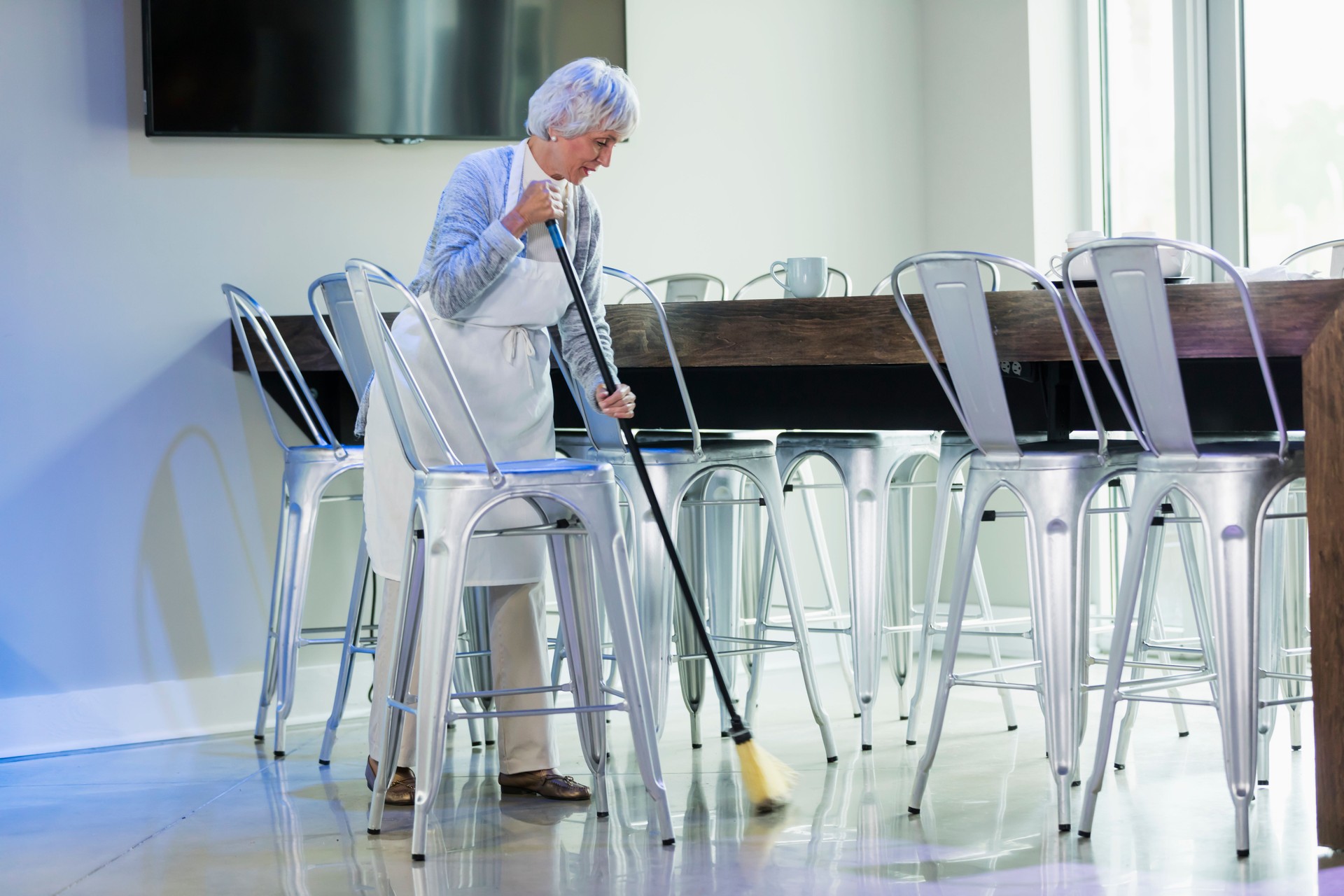 Senior woman in coffee shop tidying up, with broom